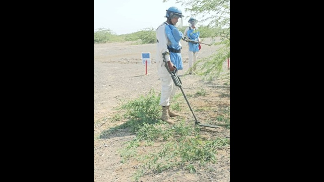 Two people wearing protective helmets, vests, and metal detectors are inspecting an open, dry outdoor area with low vegetation. Marker stakes are visible, indicating a technical work zone.; }}