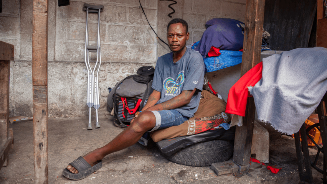 Un homme n'ayant qu'une jambe est assis sur un tas d'affaires, dans un espace encombré de tissus, sacs et autres objets. Derrière lui, appuyées contre un mur de briques, deux béquilles neuves.