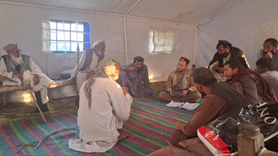 Under a canvas tent, a group of men are sitting in a circle on mats and benches. They are talking to a man wearing a jacket with HI printed on it.; }}