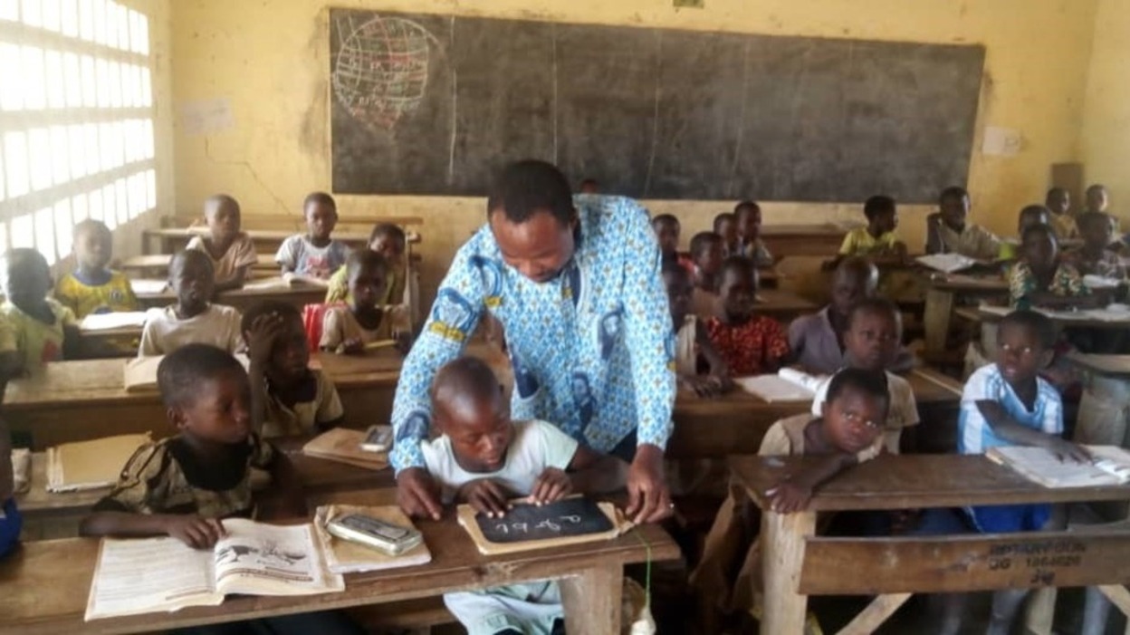 Dans une salle de classe pleine, Yao se penche au-dessus d'un enfant pour l'aider.