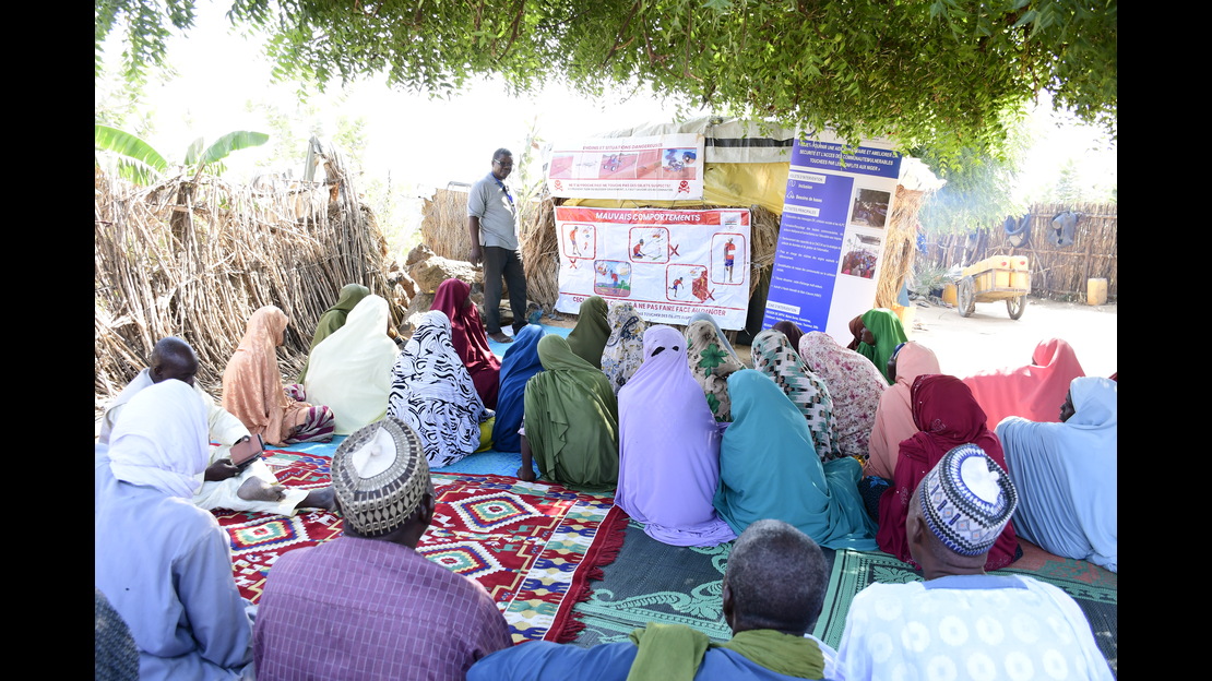  A group of people is sitting on the ground on colorful rugs and mats, arranged in the shade of a large tree in a rural setting. One person is standing in front of the group and presenting information using several posters and signs attached to a structure made of simple materials. The visual aids include illustrations, pictograms, and blocks of text. In the background, a fence made of branches surrounds the area, and a path and a cart are visible. ; }}