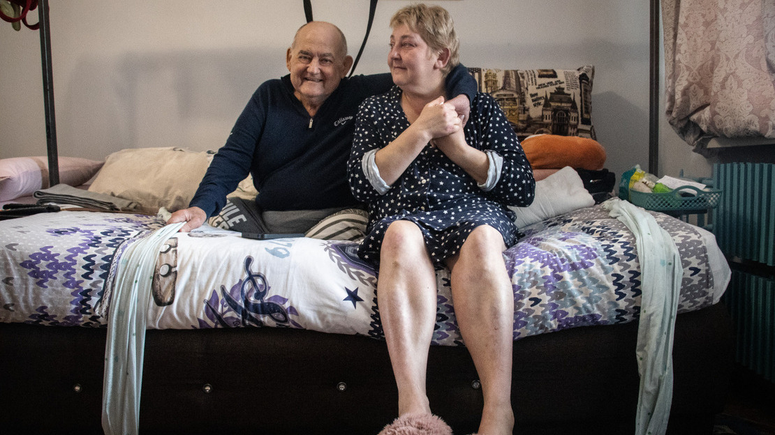 Un couple âgé est assis sur un lit, partageant un moment de complicité. L’homme, vêtu d’un pull foncé, sourit chaleureusement tandis que la femme, en robe de chambre étoilée et pantoufles roses, lui prend tendrement la main au-dessus de son épaule. L’atmosphère intime de la chambre, avec ses draps colorés et ses objets du quotidien, dégage une impression de douceur et de proximité.; }}