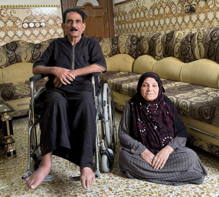 Hmoud in his living room, sitting in a wheelchair. His wife sits besides him.