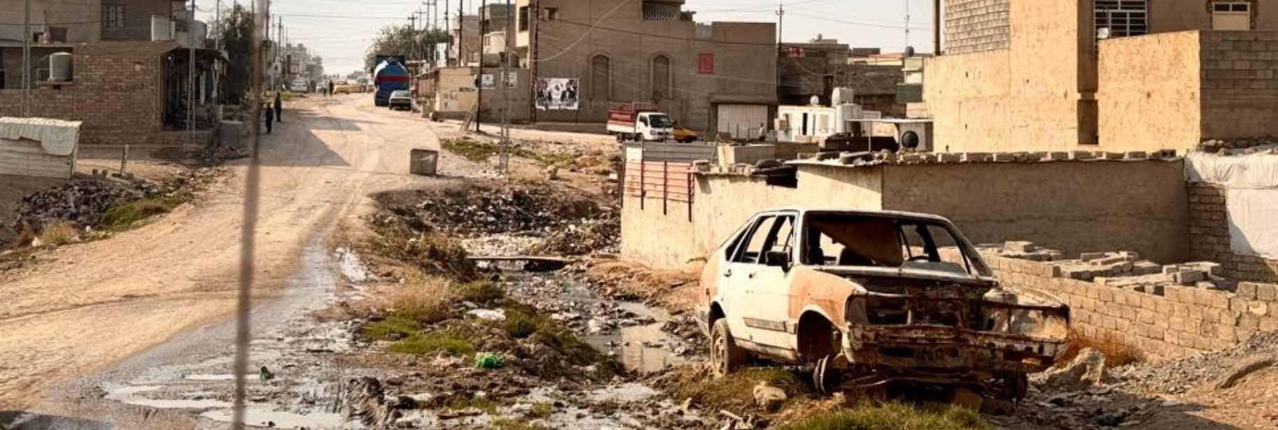 A car damaged by explosive weapons sits in a street in front of civilians’ homes.